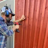 Man med hörselkåpor och keps skruvar fast ny panel utanpå den gamla röda träfasaden.