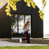 Svart trähus med stora fönster vid en sjö. En person springer förbi. Utbyggnad hus inspiration för moderna hem.