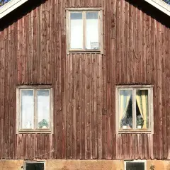 Rödbrun panel på ett gammalt timmerhus med tre fönster. Ett fönster högst upp har en skylt.