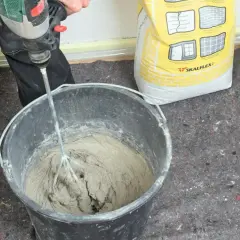En person använder en borrmaskin för att blanda betong i en hink. En gul säck med Skalflex-märkning står bredvid.