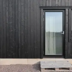 Svart husgrund med träpanel, glasdörr och fönster. En pall som trappa leder upp.