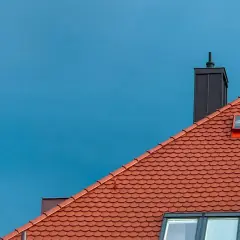 Rött tegeltak med skorsten och takfönster mot en blå himmel. Dags att byta tak?