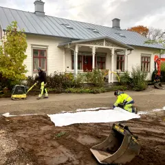 Arbetare lägger ut duk och packar grus för en gammaldags grusgång framför ett vitt hus. En grävmaskin syns i bakgrunden.