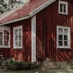Rött torp med glasveranda, en cykel och en stege står på gräsmattan.