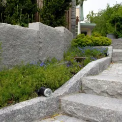 En imponerande mur av granit sten ramar in en lummig rabatt med blå blommor. En trappa i samma granit sten leder uppåt.