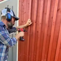 Man med hörselkåpor och keps skruvar fast ny panel utanpå den gamla röda träfasaden.