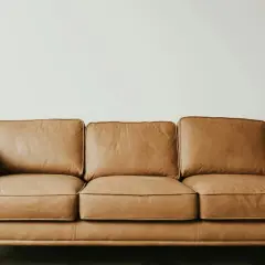 Brun lädersoffa med tre sittkuddar och ryggkuddar, på sneda träben. En grön växt syns till höger. Perfekt för hållbara hus.
