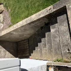 Betongtrappa leder ner till en träport. Gräs och stenar syns ovanför. Kanske någon ska borra i putsad fasad här snart.