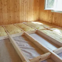 Golvbjälkar fyllda med isolering för att isolera golv ovanifrån. Träväggar och ett fönster syns i bakgrunden.
