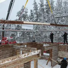 Kranen lyfter timmer för att flytta timmerhus i snöig skogsmiljö. Byggnadsarbetare övervakar arbetet.