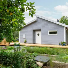Ett modernt, ljusgrått hus med träfasad och stora altaner. Trädgården är frodig med äppelträd och blommor.