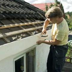 Man byter vindskivor på ett tak. Han håller en ny vit bräda och hamrar in en spik.