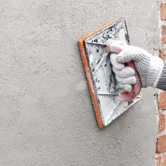 En hand med handske använder en murslev för att putsa en tegelfasad.