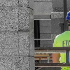 Construction worker in a bright green shirt with "FINJA" on the back, wearing a blue hard hat, next to a wall of concrete blocks.