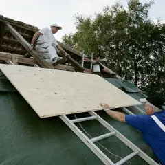 Takläggare arbetar på ett tak med råspont vilken sida upp. En stor plywoodskiva placeras på plats.