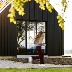 Svart trähus med stora fönster vid en sjö. En person springer förbi. Utbyggnad hus inspiration för moderna hem.