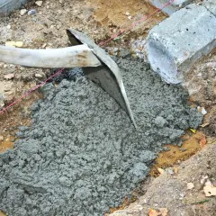En hacka sprider jordfuktig betong i en grop. Betongblock och en rosa snodd syns i bakgrunden.
