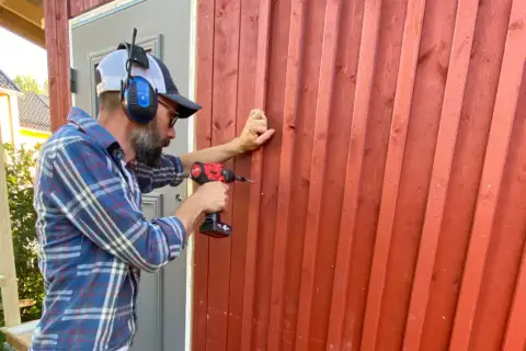 Man med hörselkåpor och keps skruvar fast ny panel utanpå den gamla röda träfasaden.