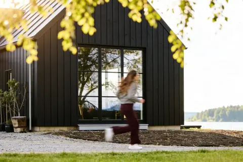 Svart trähus med stora fönster vid en sjö. En person springer förbi. Utbyggnad hus inspiration för moderna hem.