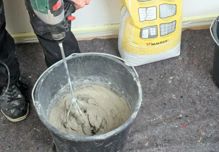 En person använder en borrmaskin för att blanda betong i en hink. En gul säck med Skalflex-märkning står bredvid.