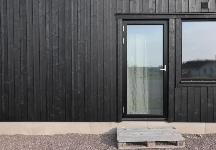 Svart husgrund med träpanel, glasdörr och fönster. En pall som trappa leder upp.