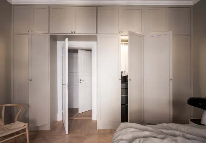 En elegant garderob i sekelskiftesstil med ljusa fronter och dörrar som leder till en walk-in closet.