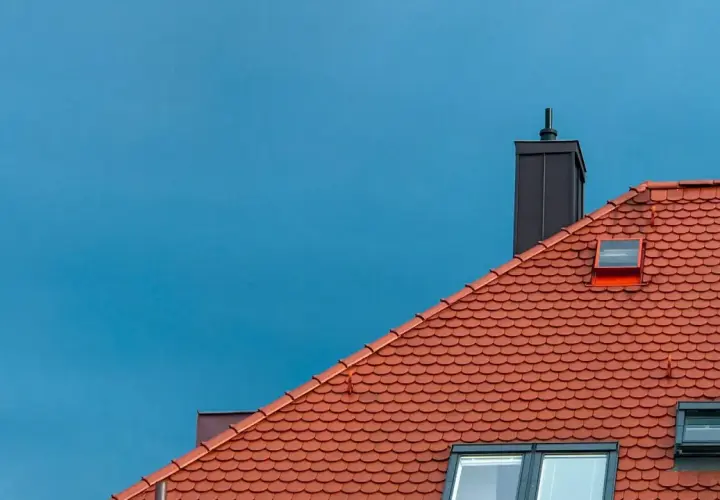 Rött tegeltak med skorsten och takfönster mot en blå himmel. Dags att byta tak?