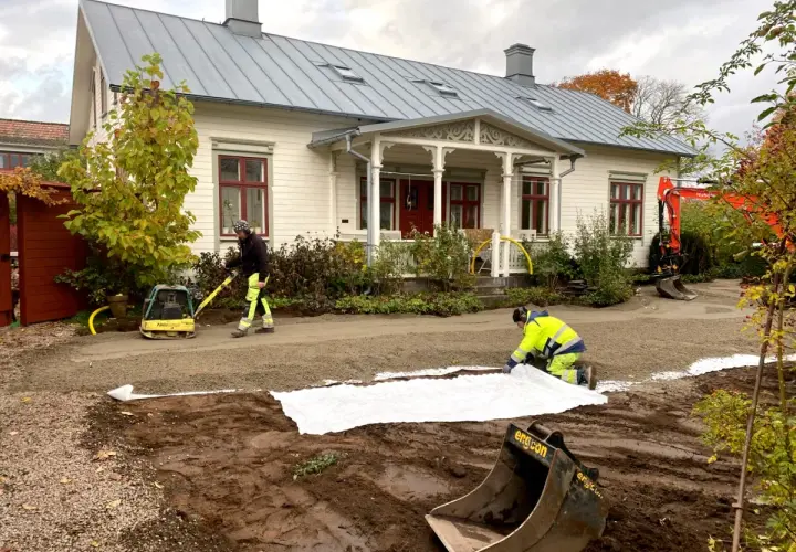 Arbetare lägger ut duk och packar grus för en gammaldags grusgång framför ett vitt hus. En grävmaskin syns i bakgrunden.