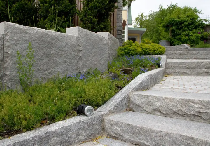 En imponerande mur av granit sten ramar in en lummig rabatt med blå blommor. En trappa i samma granit sten leder uppåt.