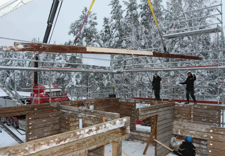 Kranen lyfter timmer för att flytta timmerhus i snöig skogsmiljö. Byggnadsarbetare övervakar arbetet.