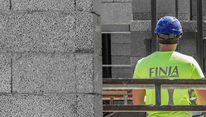 Construction worker in a bright green shirt with "FINJA" on the back, wearing a blue hard hat, next to a wall of concrete blocks.