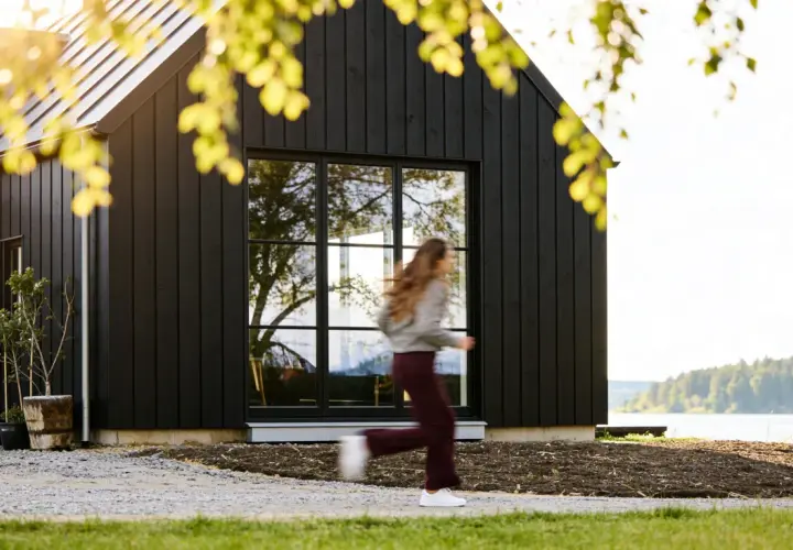 Svart trähus med stora fönster vid en sjö. En person springer förbi. Utbyggnad hus inspiration för moderna hem.