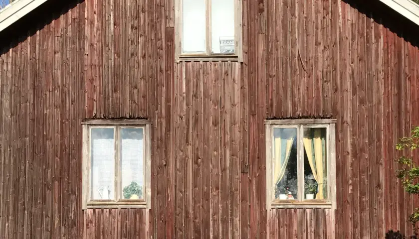Rödbrun panel på ett gammalt timmerhus med tre fönster. Ett fönster högst upp har en skylt.