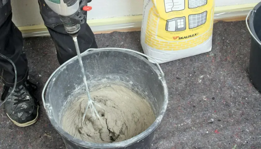En person använder en borrmaskin för att blanda betong i en hink. En gul säck med Skalflex-märkning står bredvid.