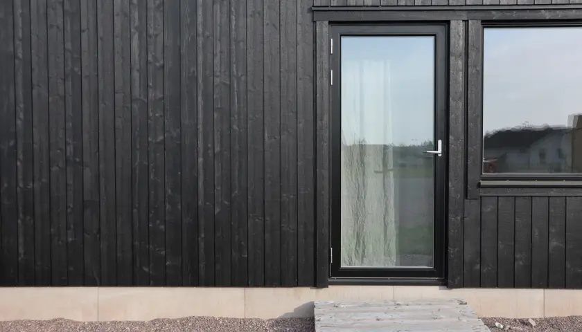 Svart husgrund med träpanel, glasdörr och fönster. En pall som trappa leder upp.