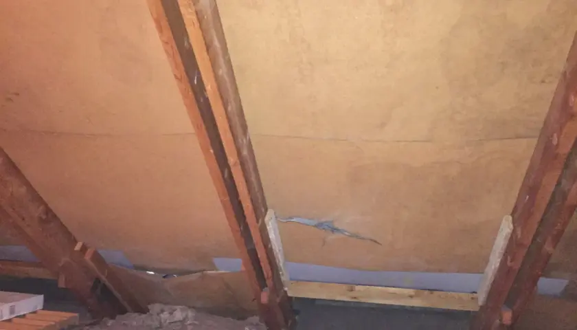 Under ett snedtak av masonit syns träbjälkar och isolering. En spricka löper genom masoniten.