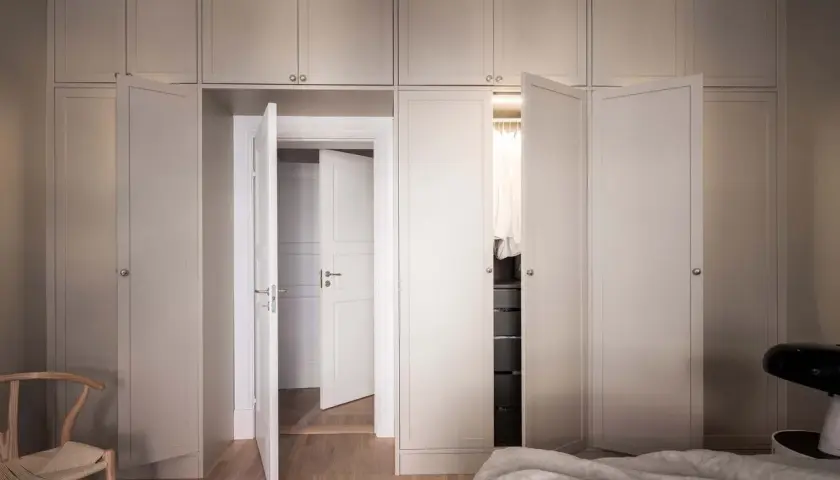 En elegant garderob i sekelskiftesstil med ljusa fronter och dörrar som leder till en walk-in closet.