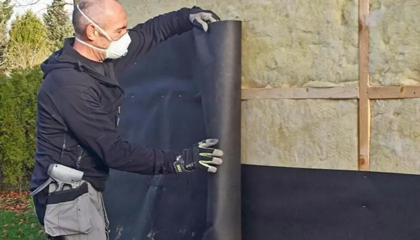 Man med mask rullar ut vindpapp på insidan av en isolerad vägg.