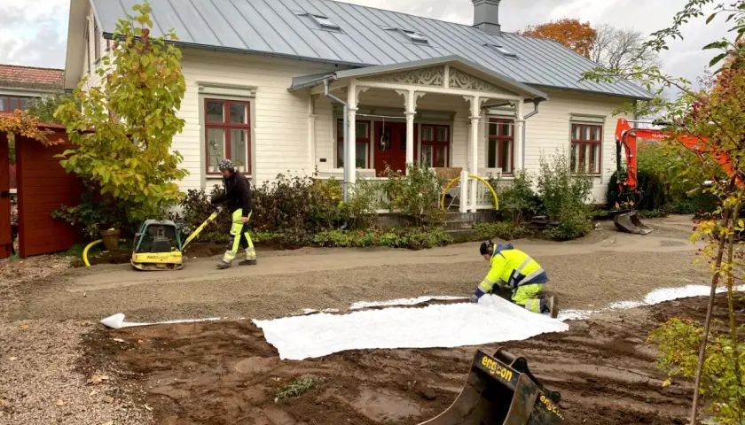 Arbetare lägger ut duk och packar grus för en gammaldags grusgång framför ett vitt hus. En grävmaskin syns i bakgrunden.