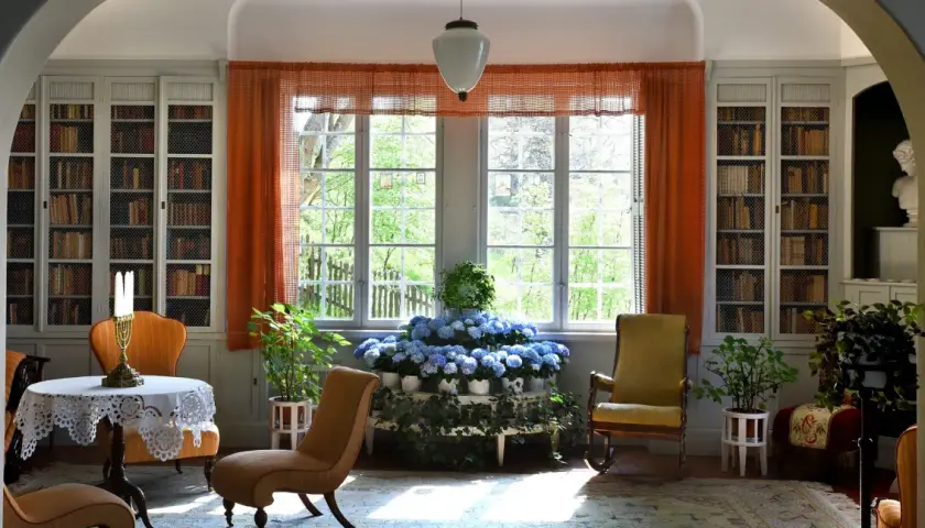 Ett rum med bokhyllor, en rund dukförsedd bord, stolar och en blomsteruppsättning. Ellen Keys inredning präglas av ljus och natur.