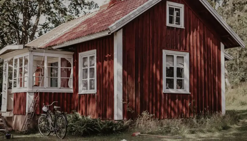 Rött torp med glasveranda, en cykel och en stege står på gräsmattan.
