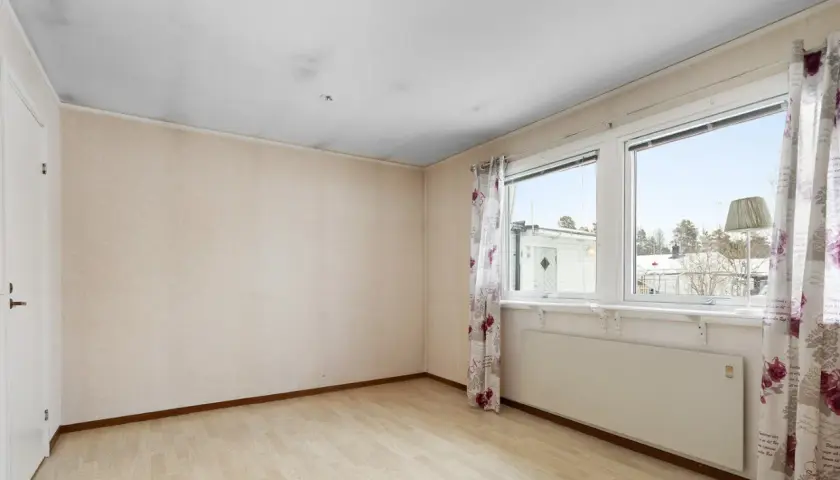Ett rum med ljusa väggar och trägolv. Svart sot i taket syns ovanför dörren och längs kanterna. Fönster med blommig gardin och utsikt över snöklädda hus.