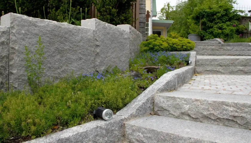 En imponerande mur av granit sten ramar in en lummig rabatt med blå blommor. En trappa i samma granit sten leder uppåt.