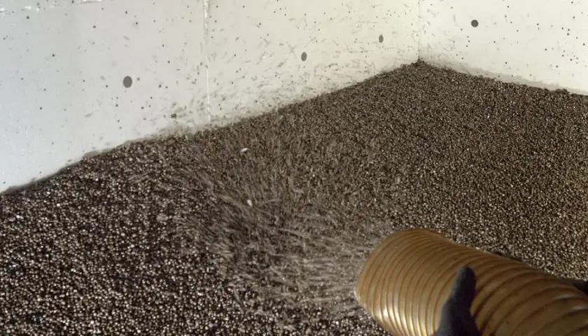 Fyller igen en torpargrund med isoleringsmaterial. En slang sprutar ut små kulor som täcker golvet.