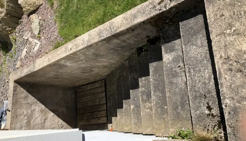 Betongtrappa leder ner till en träport. Gräs och stenar syns ovanför. Kanske någon ska borra i putsad fasad här snart.