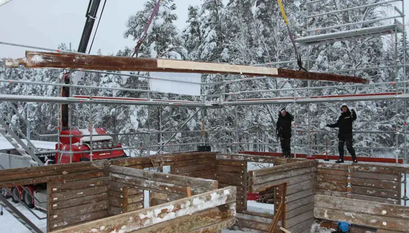 Kranen lyfter timmer för att flytta timmerhus i snöig skogsmiljö. Byggnadsarbetare övervakar arbetet.