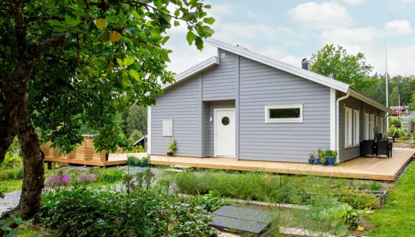 Ett modernt, ljusgrått hus med träfasad och stora altaner. Trädgården är frodig med äppelträd och blommor.