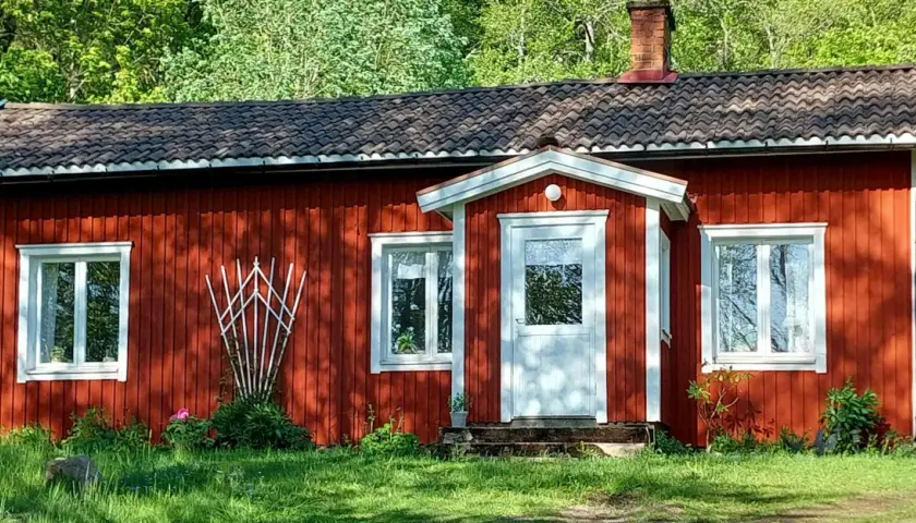 Röd stuga med vita knutar och tegeltak, omgiven av grönska. En klassisk bild som visar skillnad på torp och stuga.