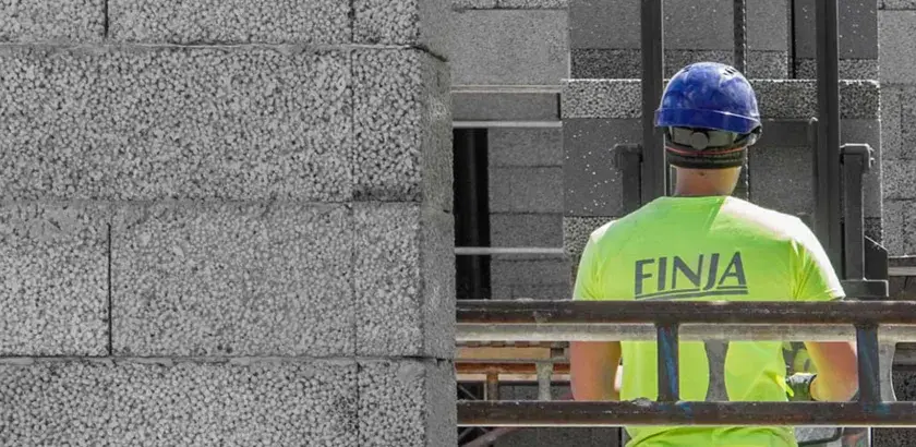 Construction worker in a bright green shirt with "FINJA" on the back, wearing a blue hard hat, next to a wall of concrete blocks.