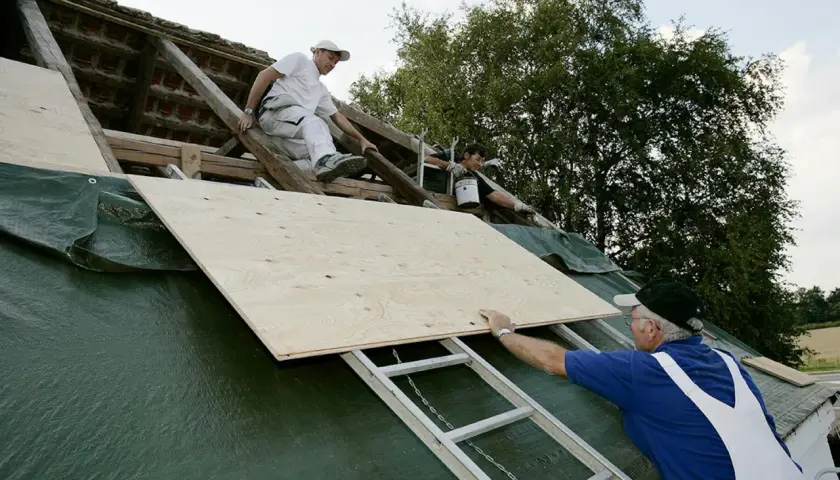 Takläggare arbetar på ett tak med råspont vilken sida upp. En stor plywoodskiva placeras på plats.